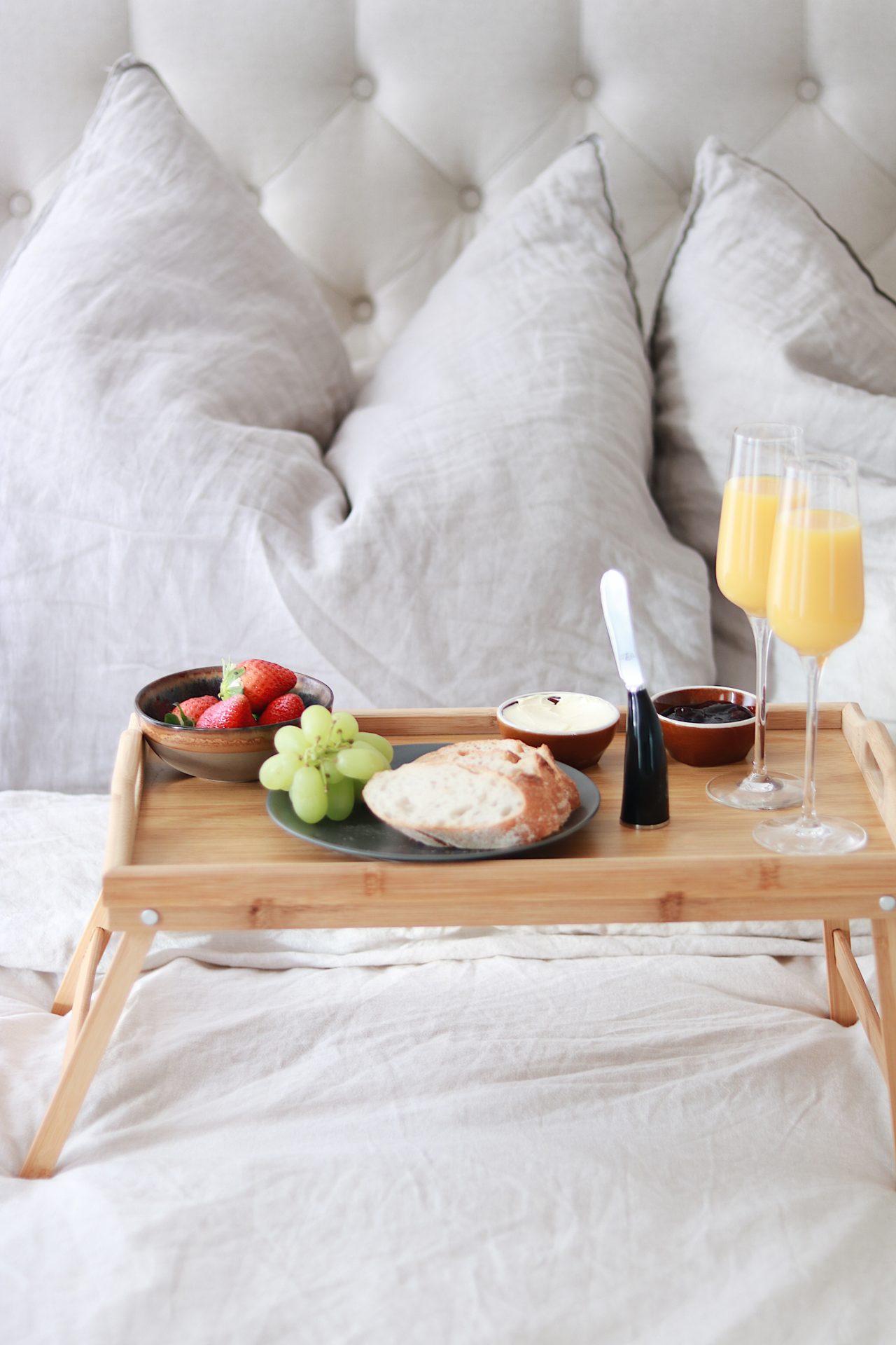 Ein Holztablett steht auf einem Bett vor beigen Kissen und ist mit Brot, Trauben, Erdbeeren, Aufstrichen und zwei Gläsern Orangensaft serviert.