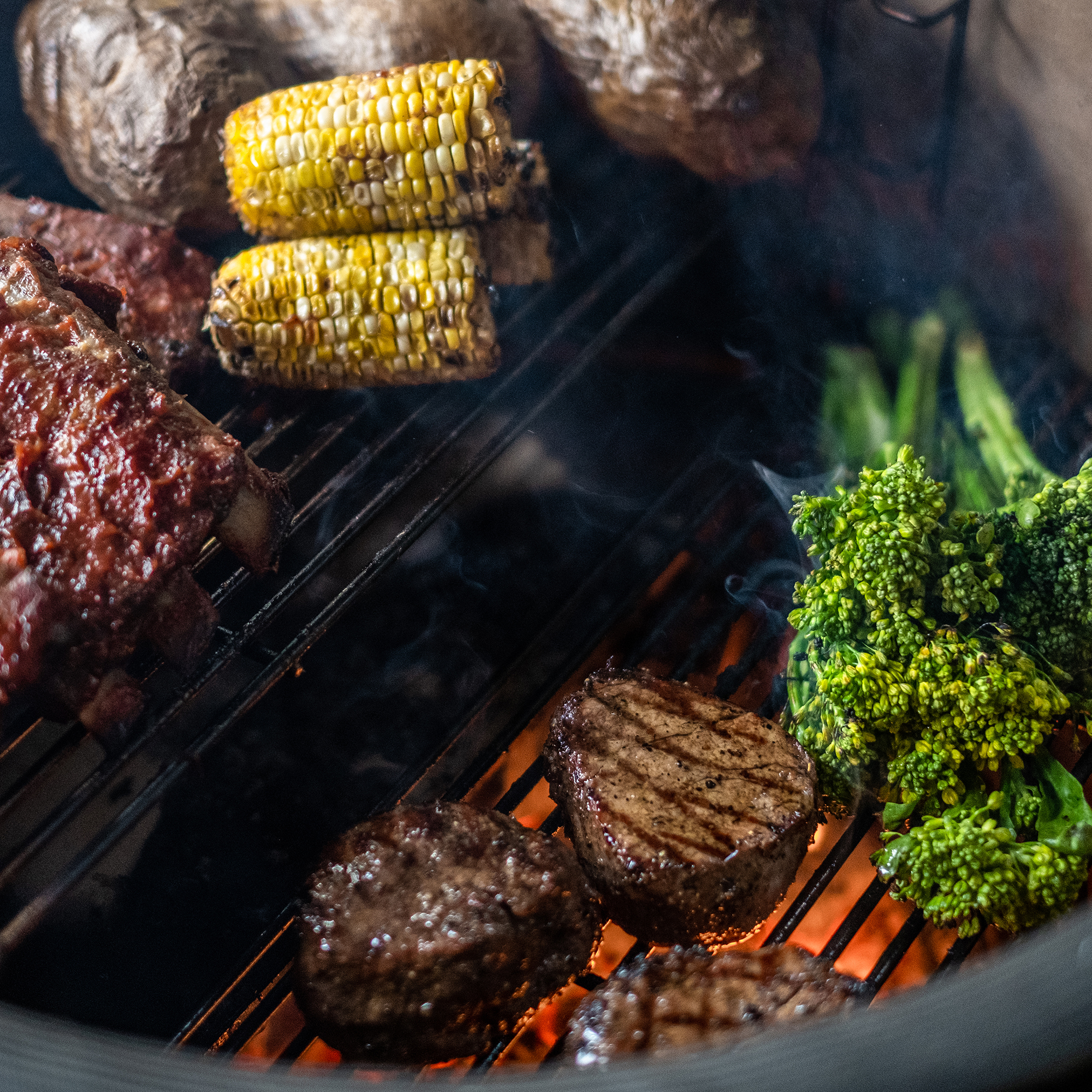 Verschiedene Speisen wie Rindersteaks, Spareribs, Maiskolben, Brokkoli und Kartoffeln werden frisch auf einem Grill zubereitet