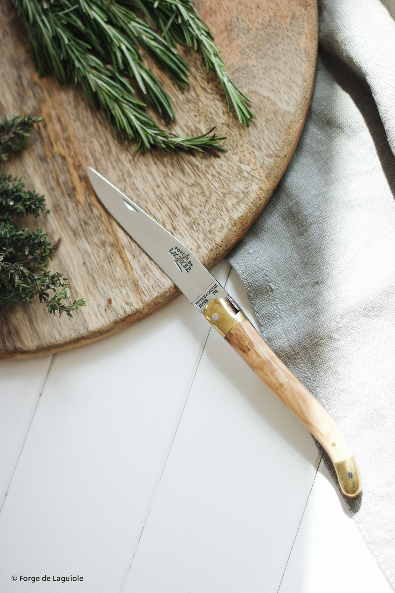 Elegantes Laguiole Messer mit Holzgriff liegt auf einem weißen Tisch neben Kräutern und einem Holzbrett