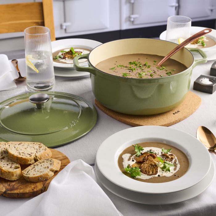 Gedeckter Tisch mit grüner Gusseisen-Kasserolle, cremiger Pilzsuppe in Tellern, frischem Brot und Wassergläsern