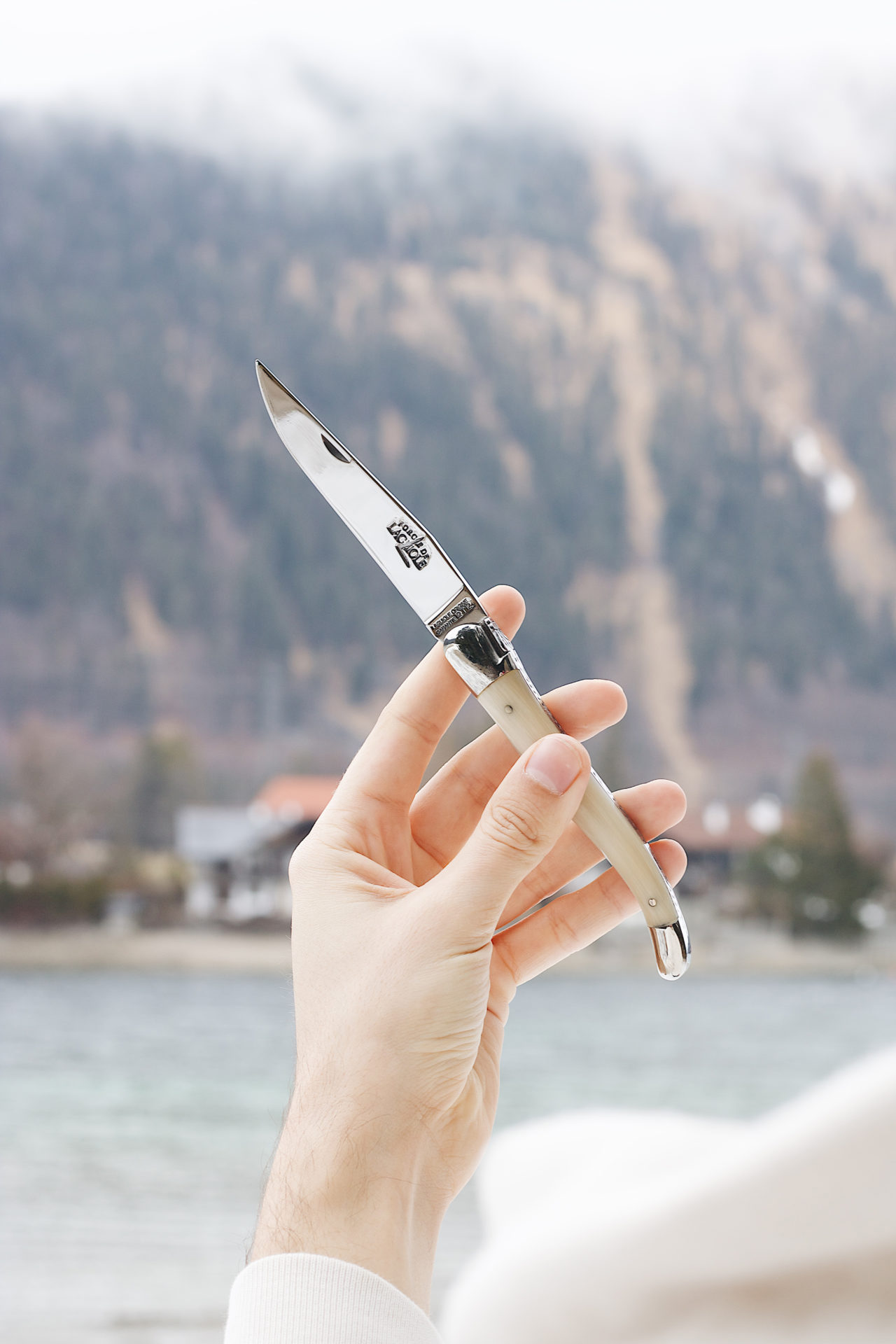 Hand hält ein elegantes Taschenmesser mit silberner Klinge und hellen Griff vor verschwommenem Gebirgshintergrund