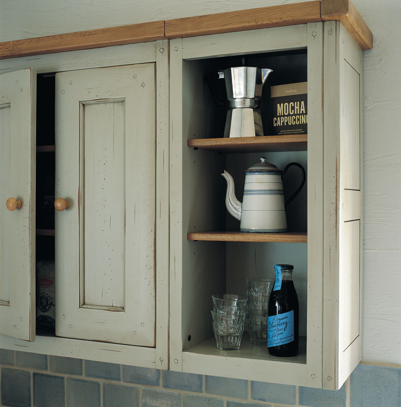 Geöffneter Landhaus-Küchenschrank mit einer silbernen Espressokanne, Mocha Cappuccino Box, Emaillekanne, vier Gläsern und einer Flasche Heidelbeersaft