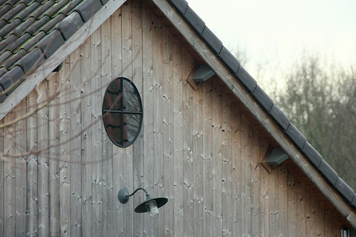 Giebel eines Hauses aus Holz mit rundem Fenster und einer schwarzen Außenlampe darunter