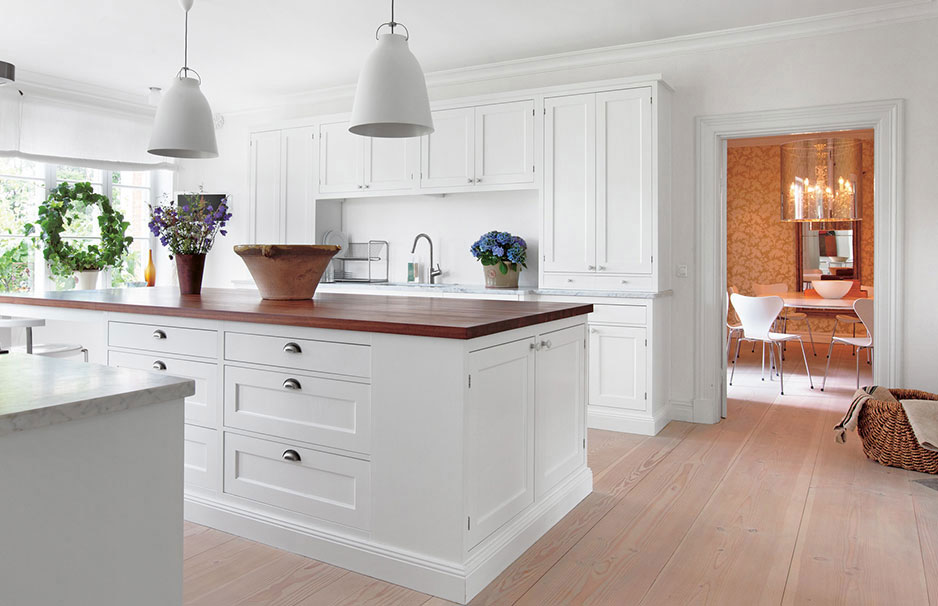 Helle, moderne Landhausküche mit weißer Kücheninsel, Holzarbeitsplatte, Hängelampen und Pflanzen. Im Hintergrund Esszimmer mit Kronleuchter sichtbar.