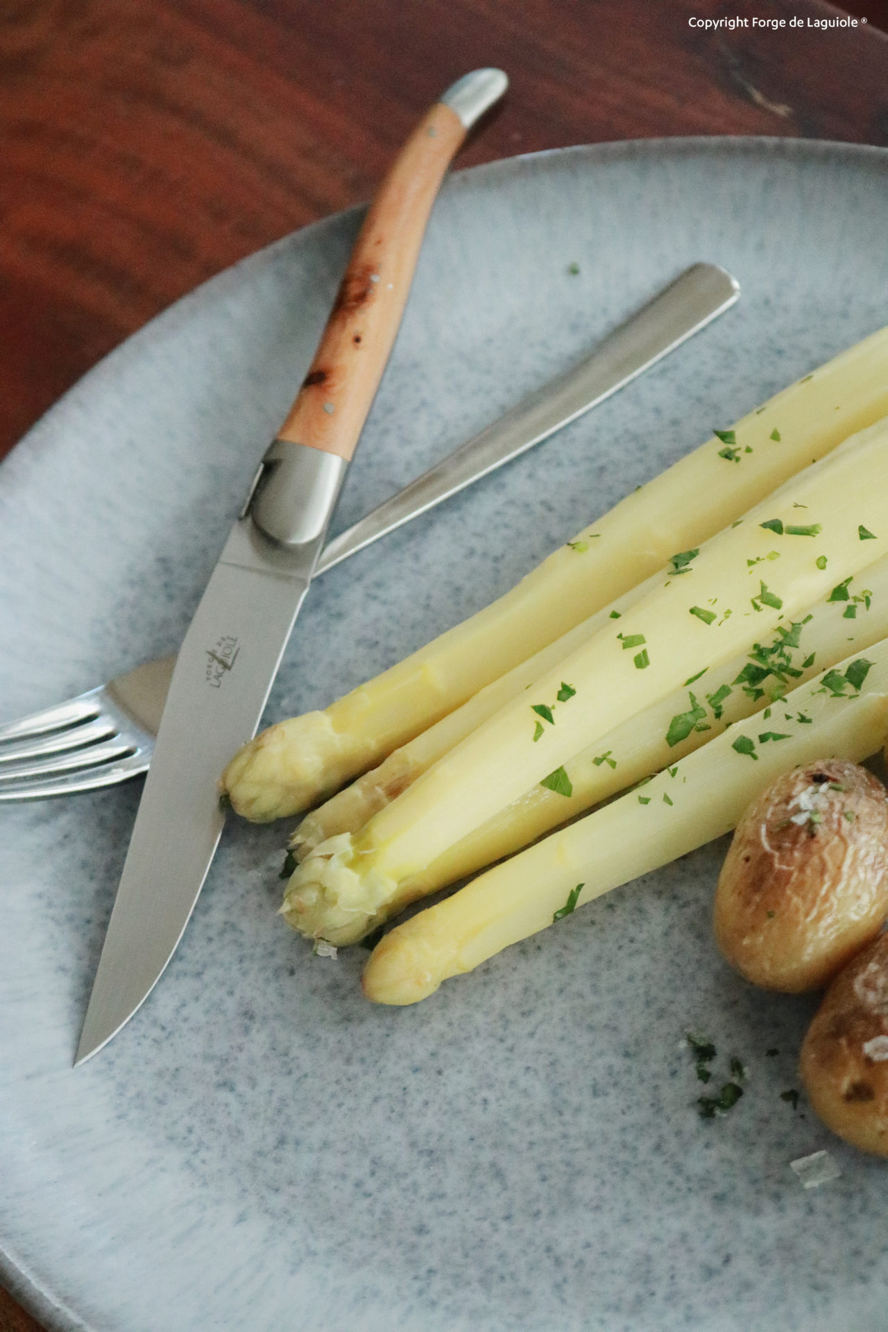 Nahaufnahme eines Tellers mit weißem Spargel, kleinen Kartoffeln und einem Messer mit Holzgriff sowie Gabel