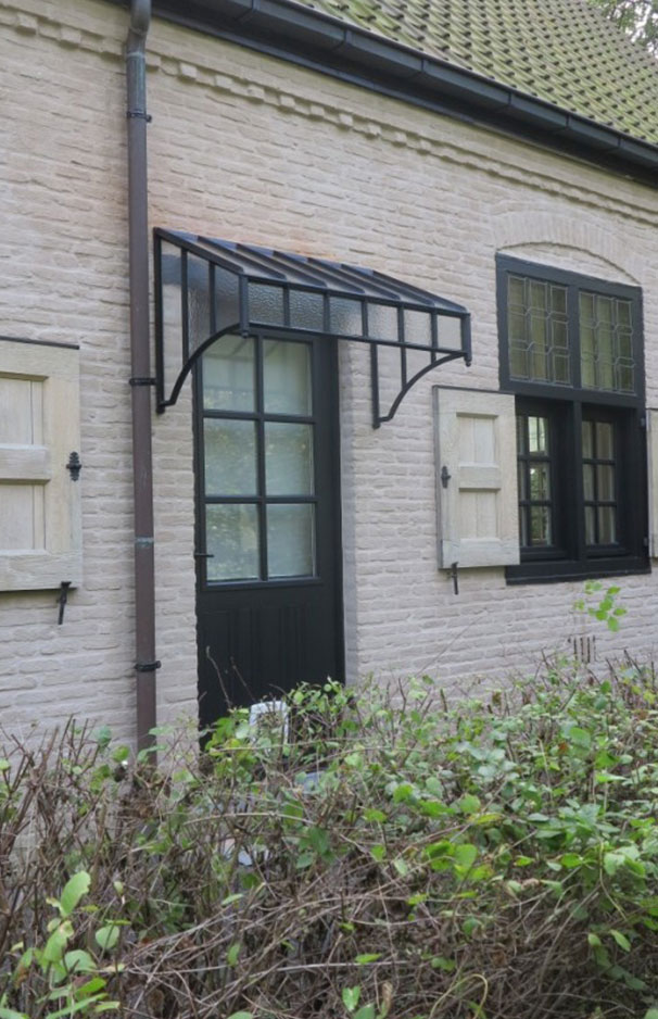 Modernes Vordach aus schwarzem Metall und Glas über einer dunklen Haustür in einer Backsteinwand mit weißen Fensterläden