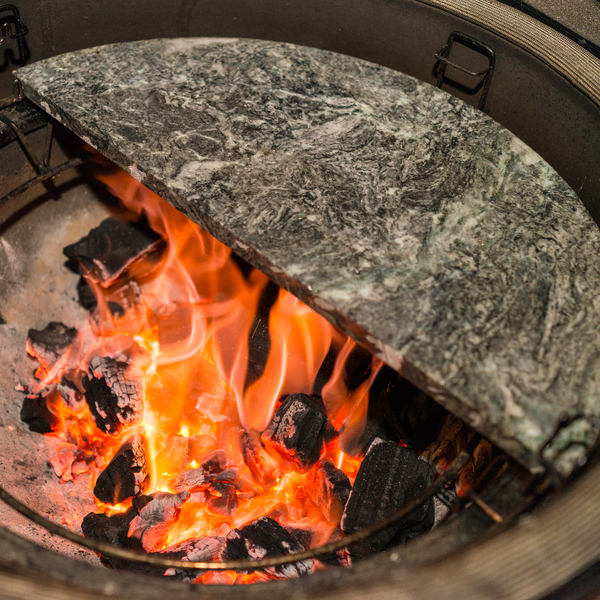 Ein halbrunder Pizzastein aus Naturstein liegt im Grill direkt über brennender Holzkohle mit Flammen.