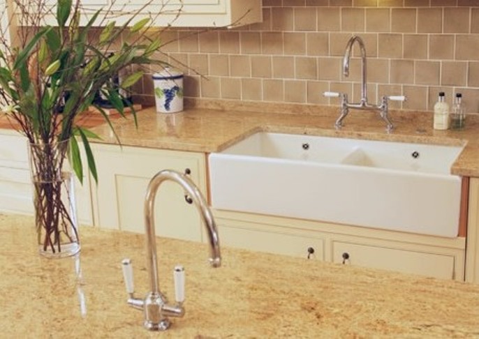 Große weiße Keramikspüle mit Doppelarmatur in cremefarbener Landhausküche, beige Fliesenrückwand, Blumenstrauß und Küchenutensilien auf der Arbeitsplatte