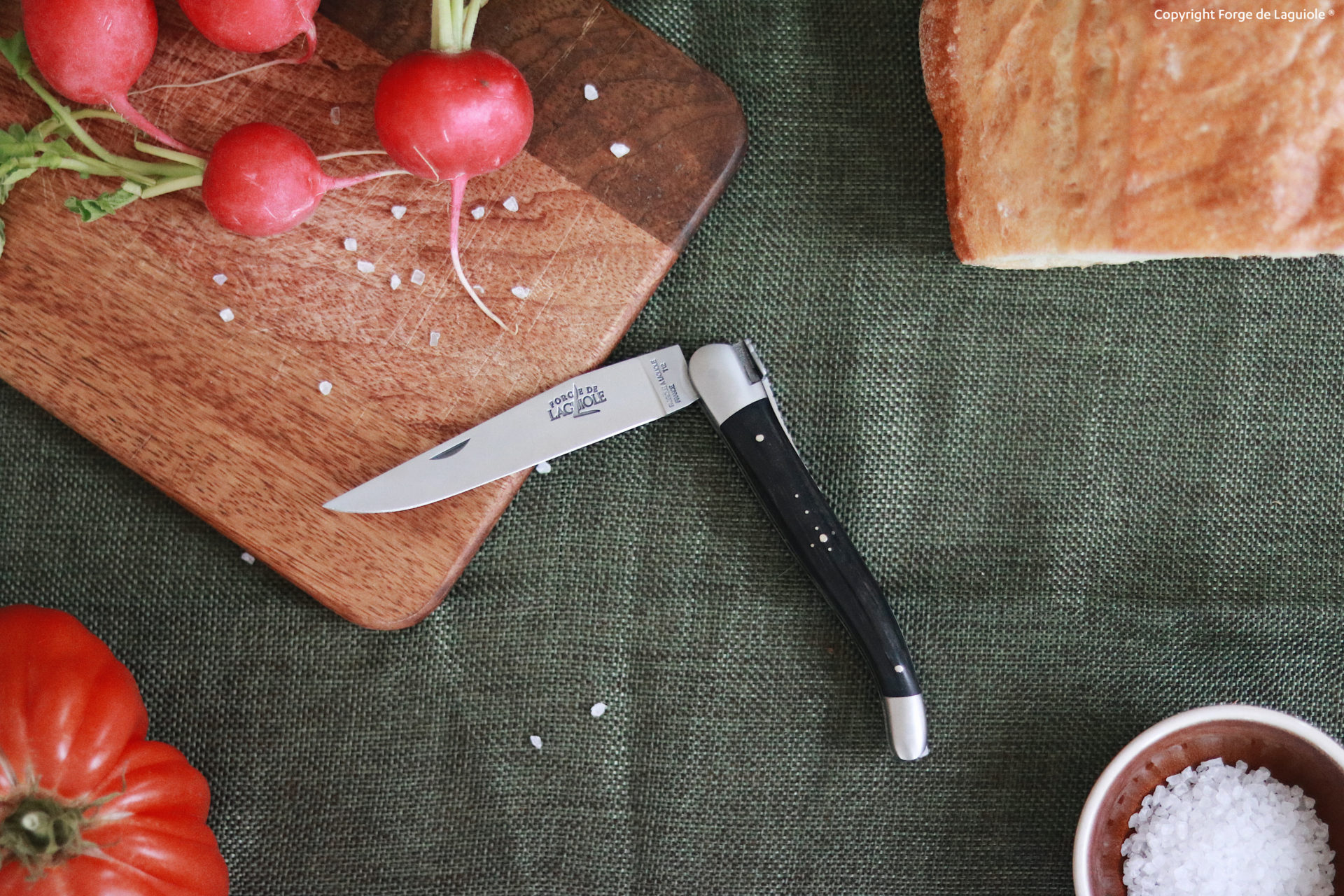 Laguiole Taschenmesser mit schwarzem Griff liegt auf einem grünen Tuch neben Schneidebrett mit Radieschen, einer Tomate, einem Laib Brot und einer Schale Meersalz