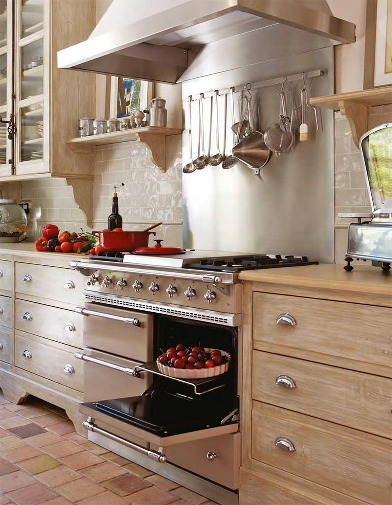 Küche im Landhausstil mit Edelstahlherd, Küchenutensilien, frischem Gemüse auf der Arbeitsplatte und offenem Backofen mit Tomaten.