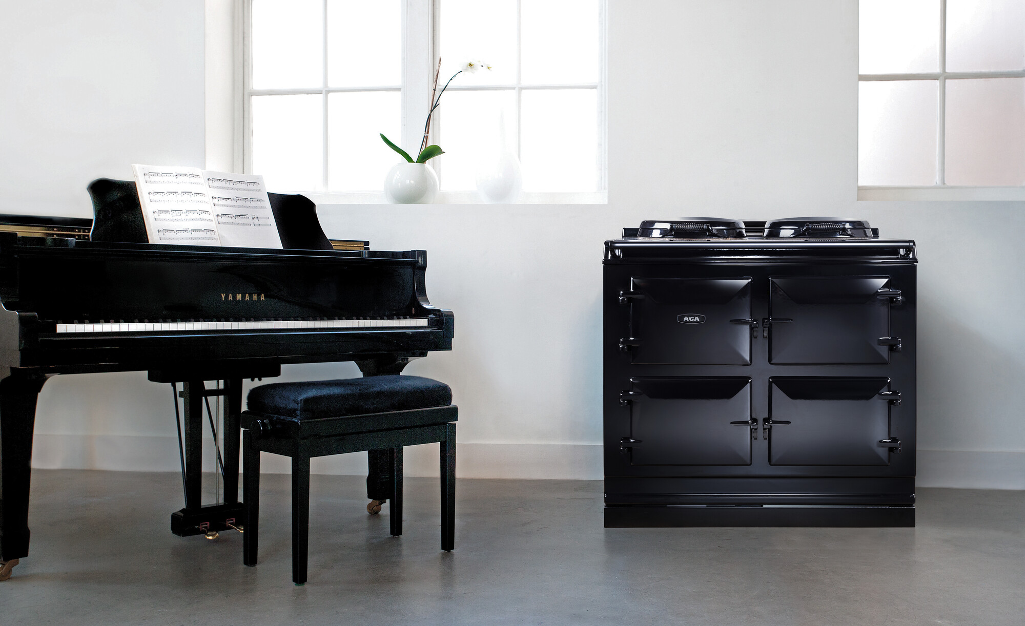 Ein schwarzer Yamaha Flügel mit offenem Notenblatt steht neben einem schwarzen AGA Herd in einem hellen, minimalistischen Raum mit weißen Wänden und zwei Fenstern im Hintergrund.