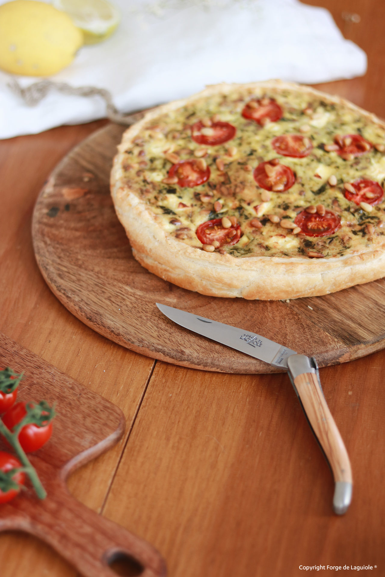 Frische Quiche mit Tomaten und Pinienkernen auf einem runden Holzbrett, daneben ein Messer und Kirschtomaten auf einem weiteren Brett
