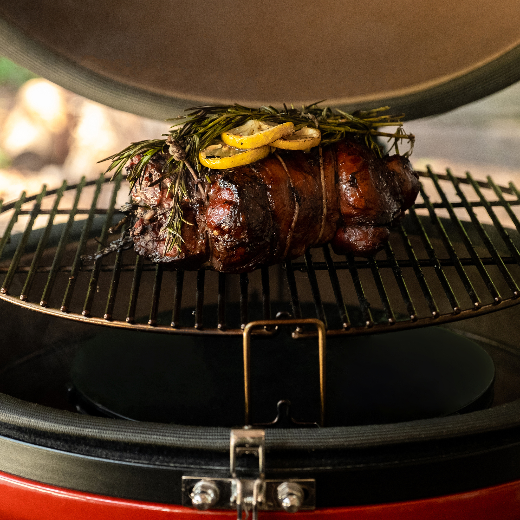 Bratenstück mit Rosmarin und Zitronenscheiben auf einem Grillrost in einem runden Keramikgrill
