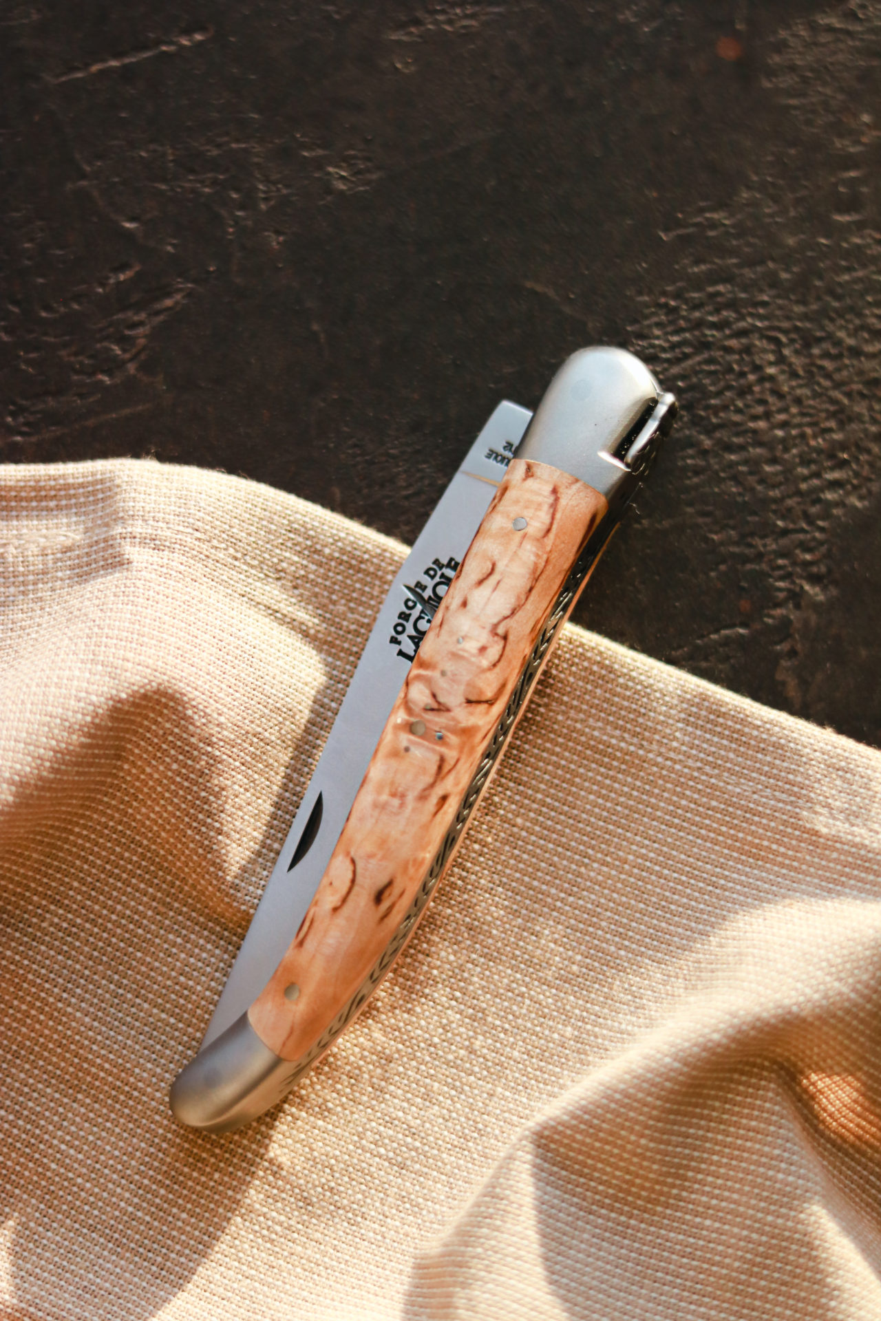 Taschenmesser mit elegant gemasertem Holzgriff und silberner Edelstahlklinge auf beigem Stoff