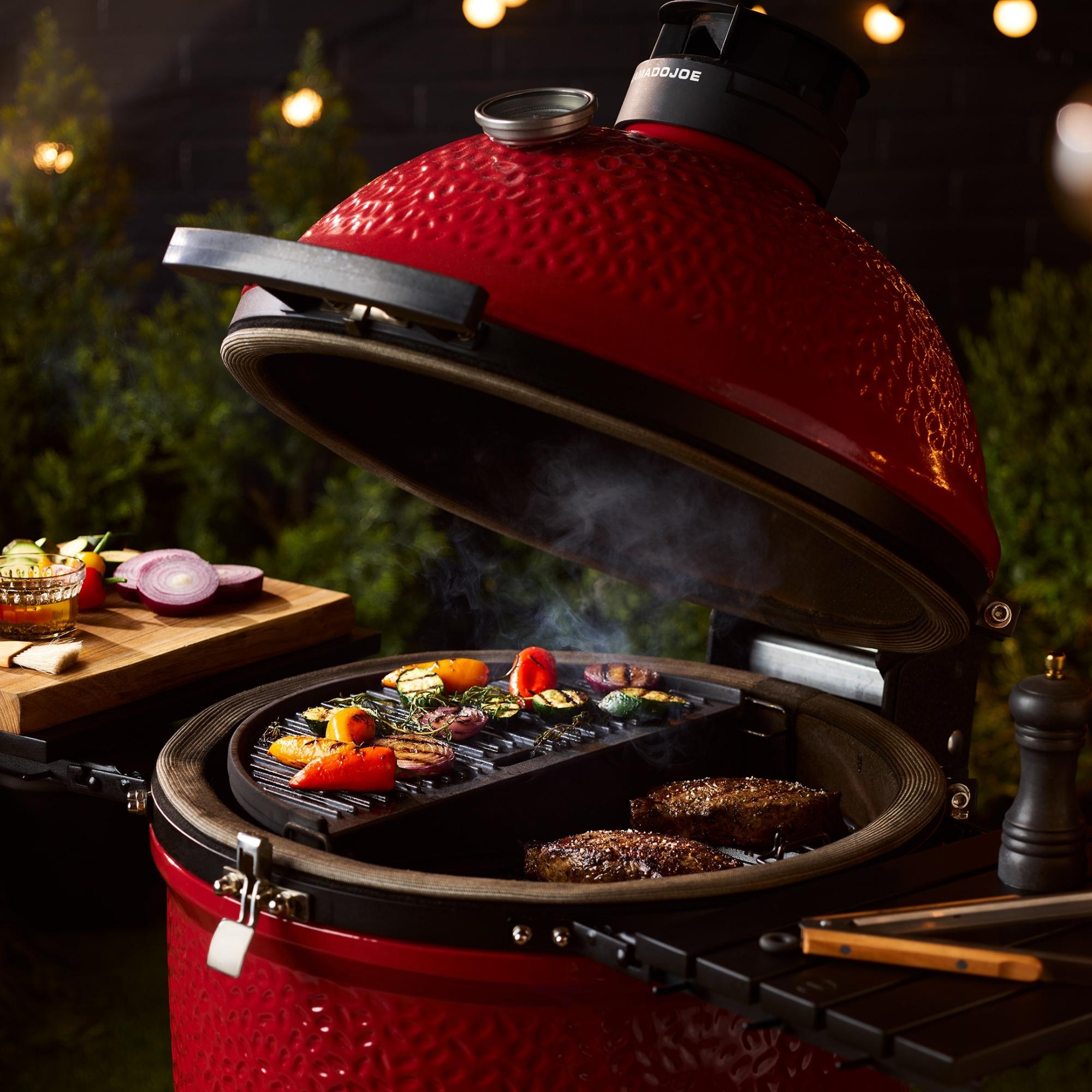 Offener roter Kamado Joe Keramikgrill mit gegrilltem Gemüse und Fleisch, daneben Schneidebrett mit Zwiebeln, draußen bei Abendbeleuchtung