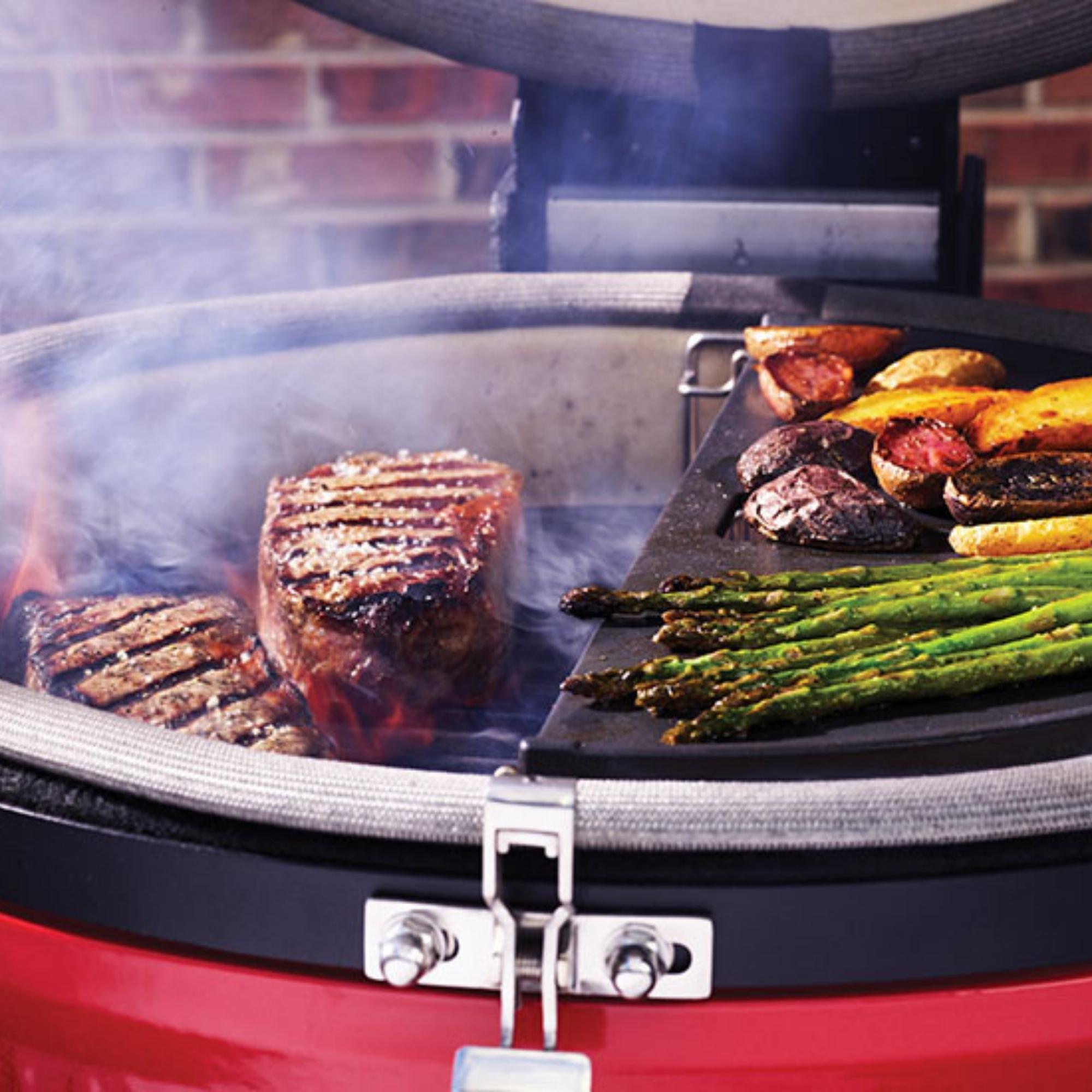 Saftige Steaks, grüner Spargel und Kartoffeln werden nebeneinander auf einem modernen Grill mit Grillplatte zubereitet, im Hintergrund ist Rauch zu sehen.