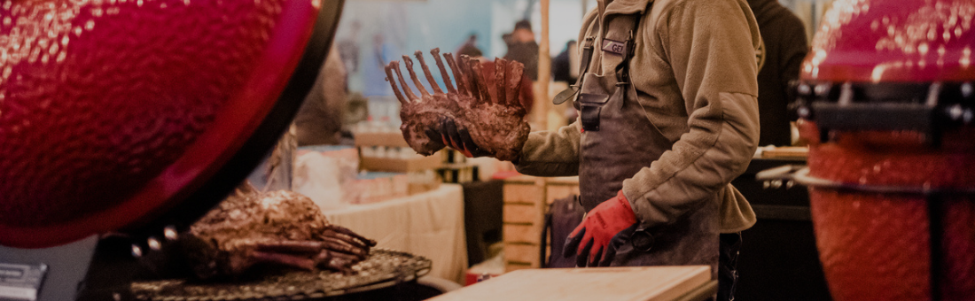 Eine Person mit Schürze und roten Handschuhen legt ein Lammkarree auf einen Grill mit rotem Keramikdeckel auf einem Markt