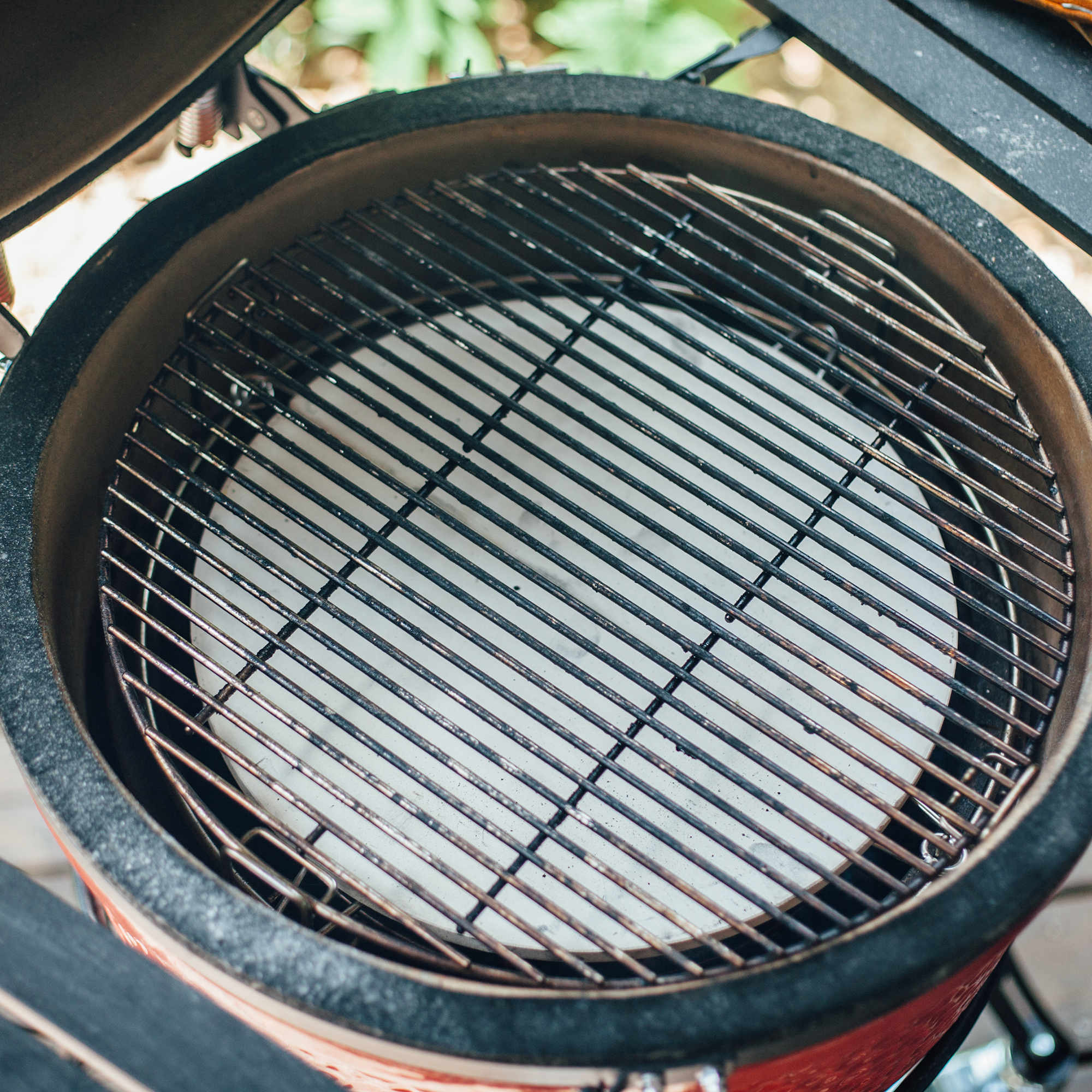 Offener Keramikgrill mit Metall-Grillrost und rundem Pizzastein, ideal für indirektes Grillen und Backen im Garten