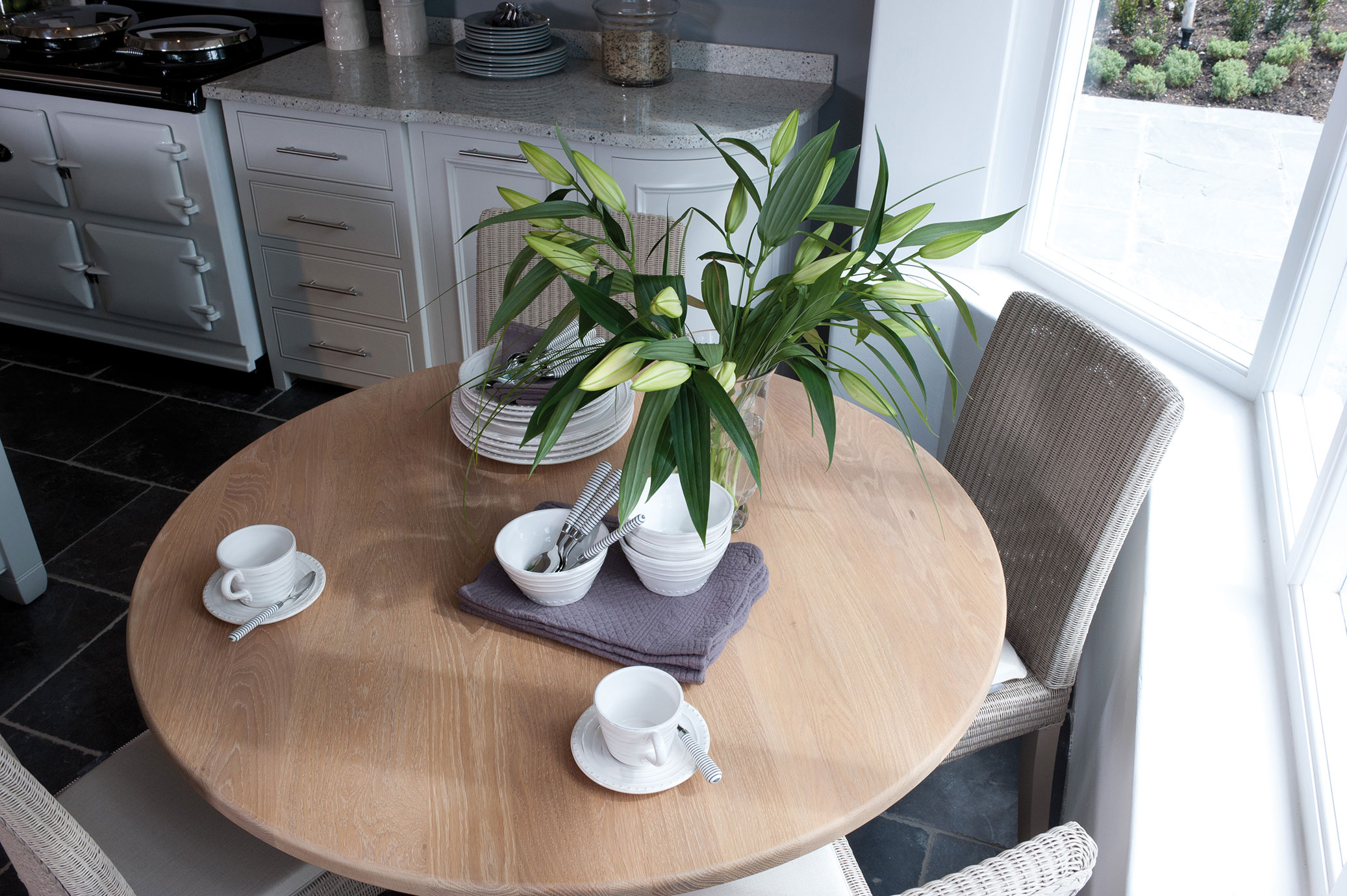 Ein runder Holztisch in einer modernen Küche ist mit weißen Keramiktassen, Untertassen, Schalen, Besteck und einer großen Vase mit grünen Blumen gedeckt. Im Hintergrund sind helle Küchenmöbel und ein Herd zu sehen.