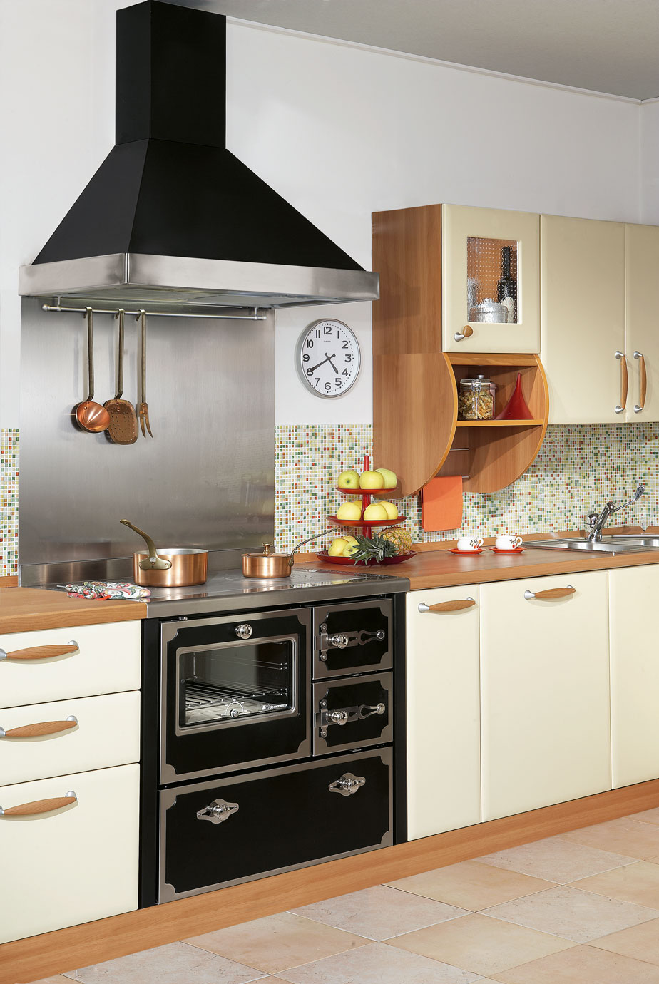 Küchenzeile mit cremefarbenen Schränken, nostalgischem Herd, Dunstabzugshaube, Kupfertöpfen, Obst-Etagere und Wanduhr vor bunter Mosaik-Fliese.