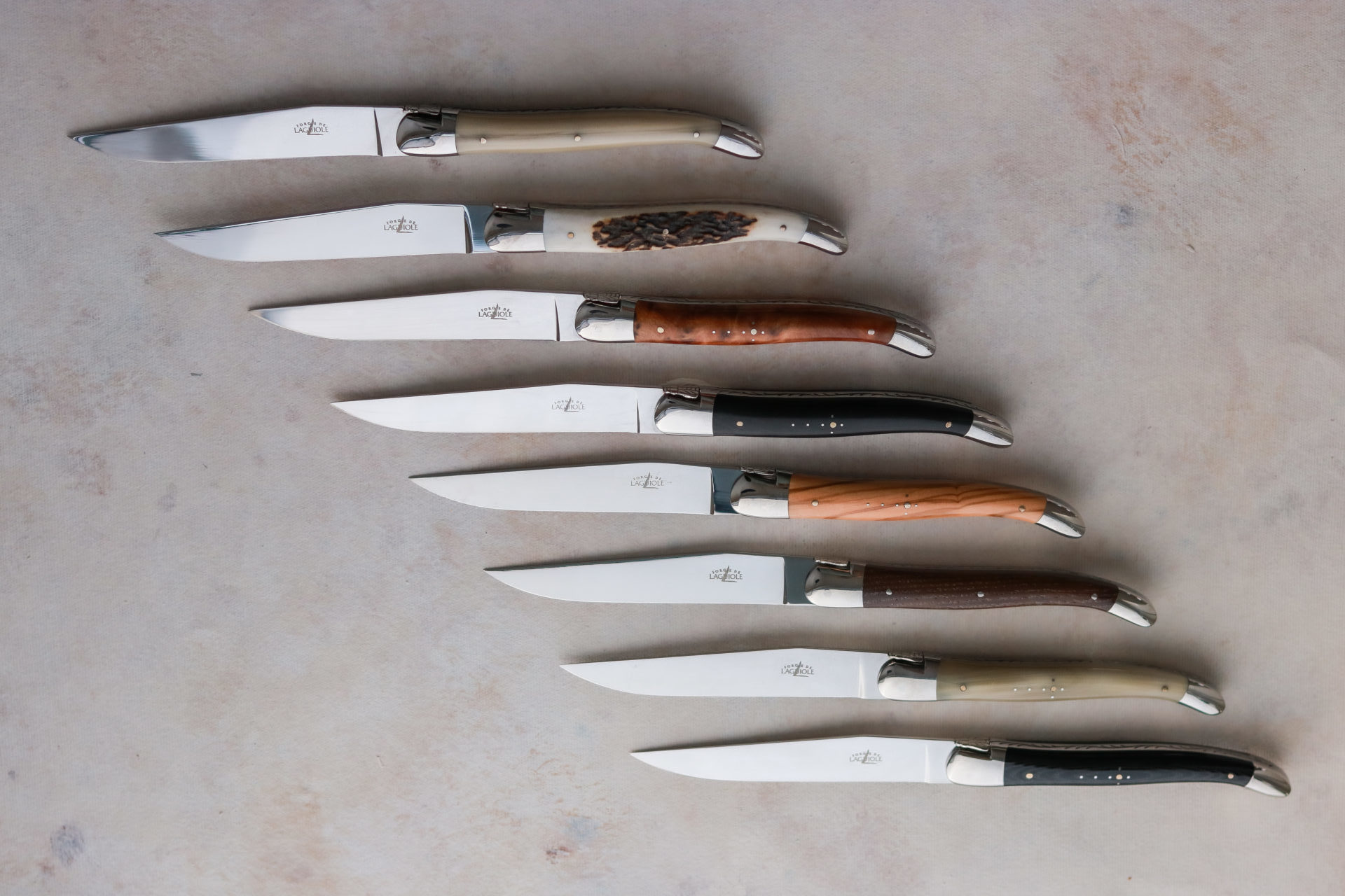 Acht verschiedene Laguiole Steakmesser mit Edelstahlklingen und Griffen aus unterschiedlichen Materialien, auf einer hellen Oberfläche angeordnet