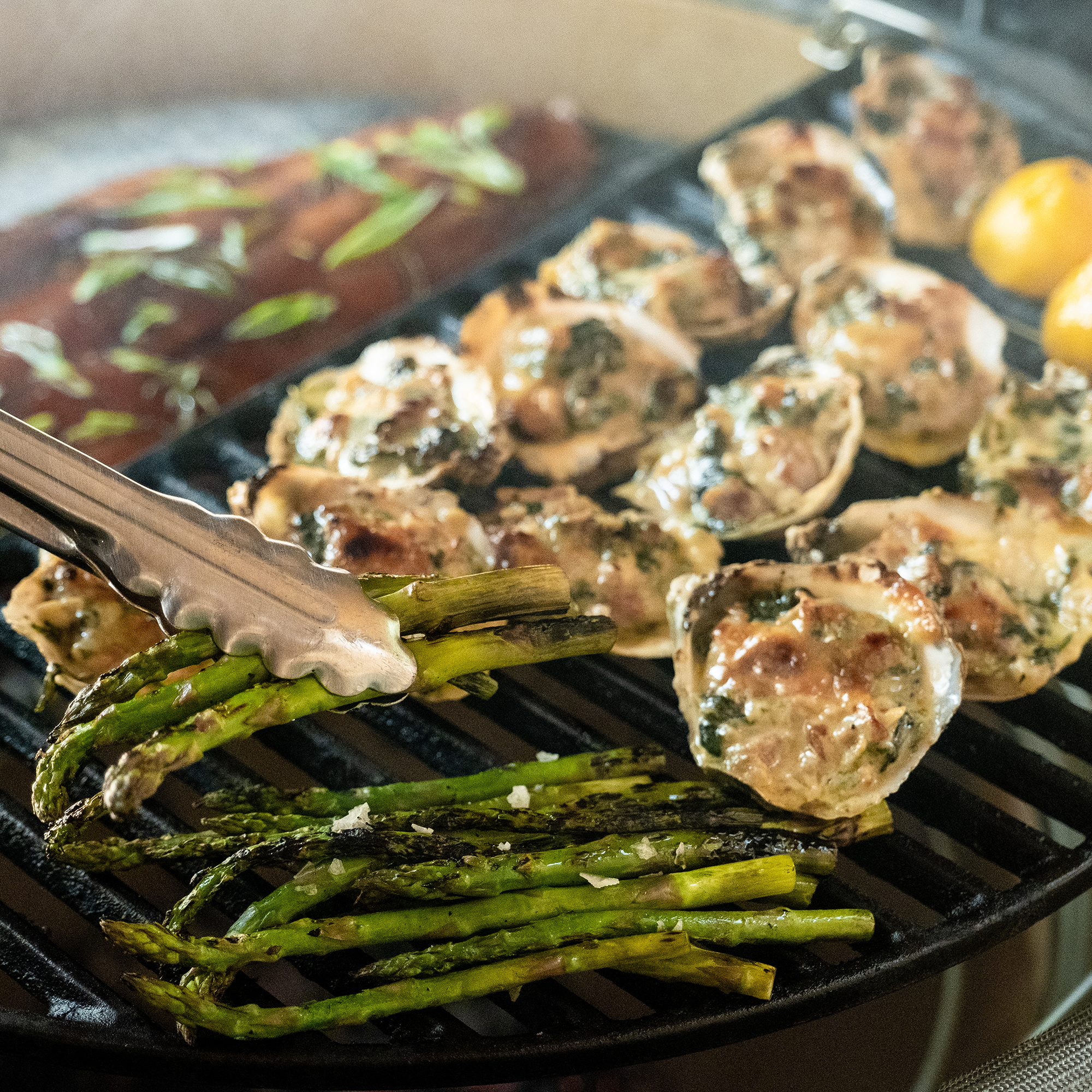 Zange hält gegrillten grünen Spargel über Grillrost, daneben gratinierte Muscheln und Zitronen