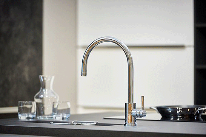 Glänzende Küchenarmatur aus Edelstahl an einem Spülbecken, im Hintergrund Wasserkrug, Gläser und Pfanne in moderner Küche