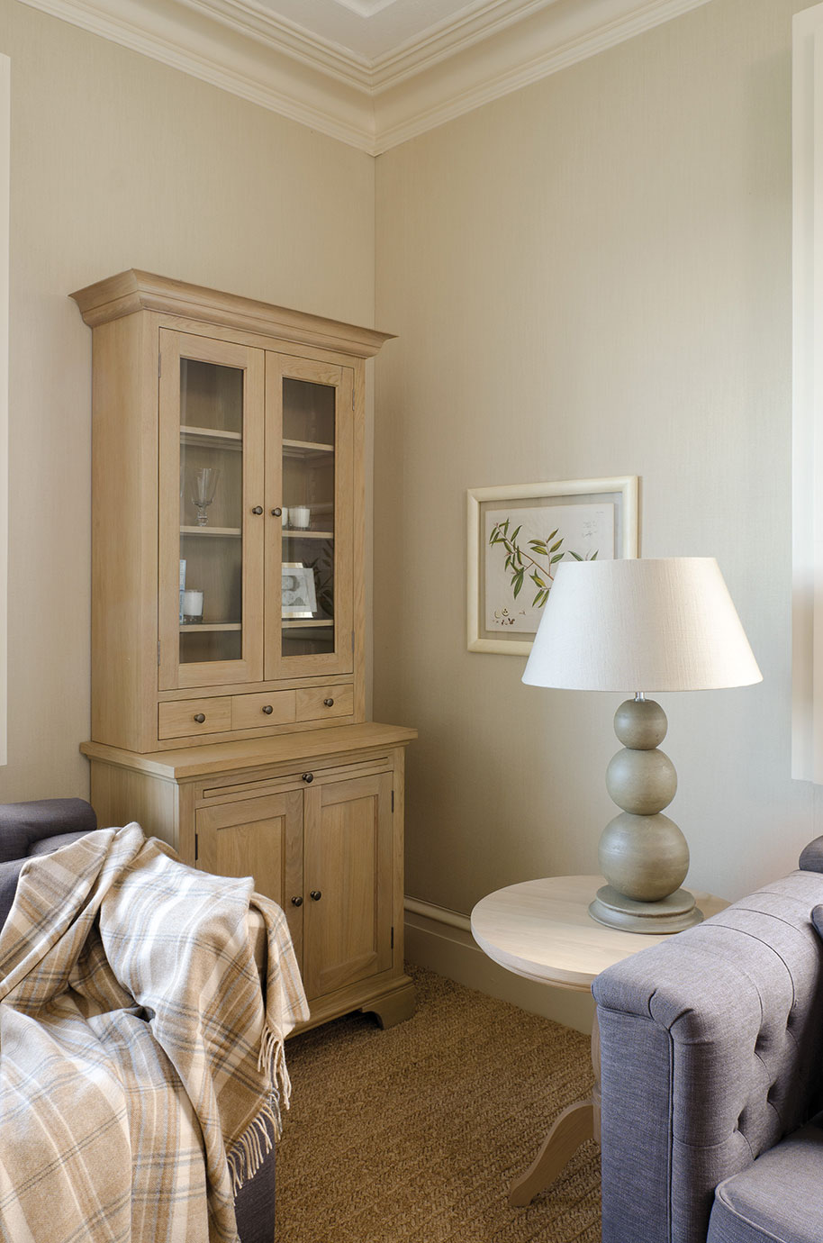 Wohnzimmer mit heller Holzvitrine, rundem Beistelltisch, Keramik-Tischlampe, grauem Sofa und beiger Decke