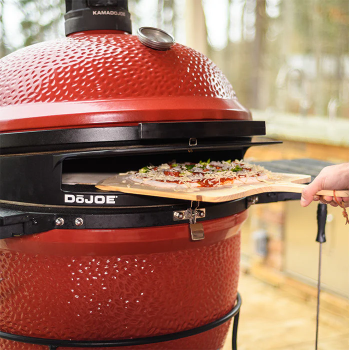 Frische Pizza auf Holzschieber wird in einen roten Kamado Joe Grill mit DoJoe Pizzaeinsatz geschoben
