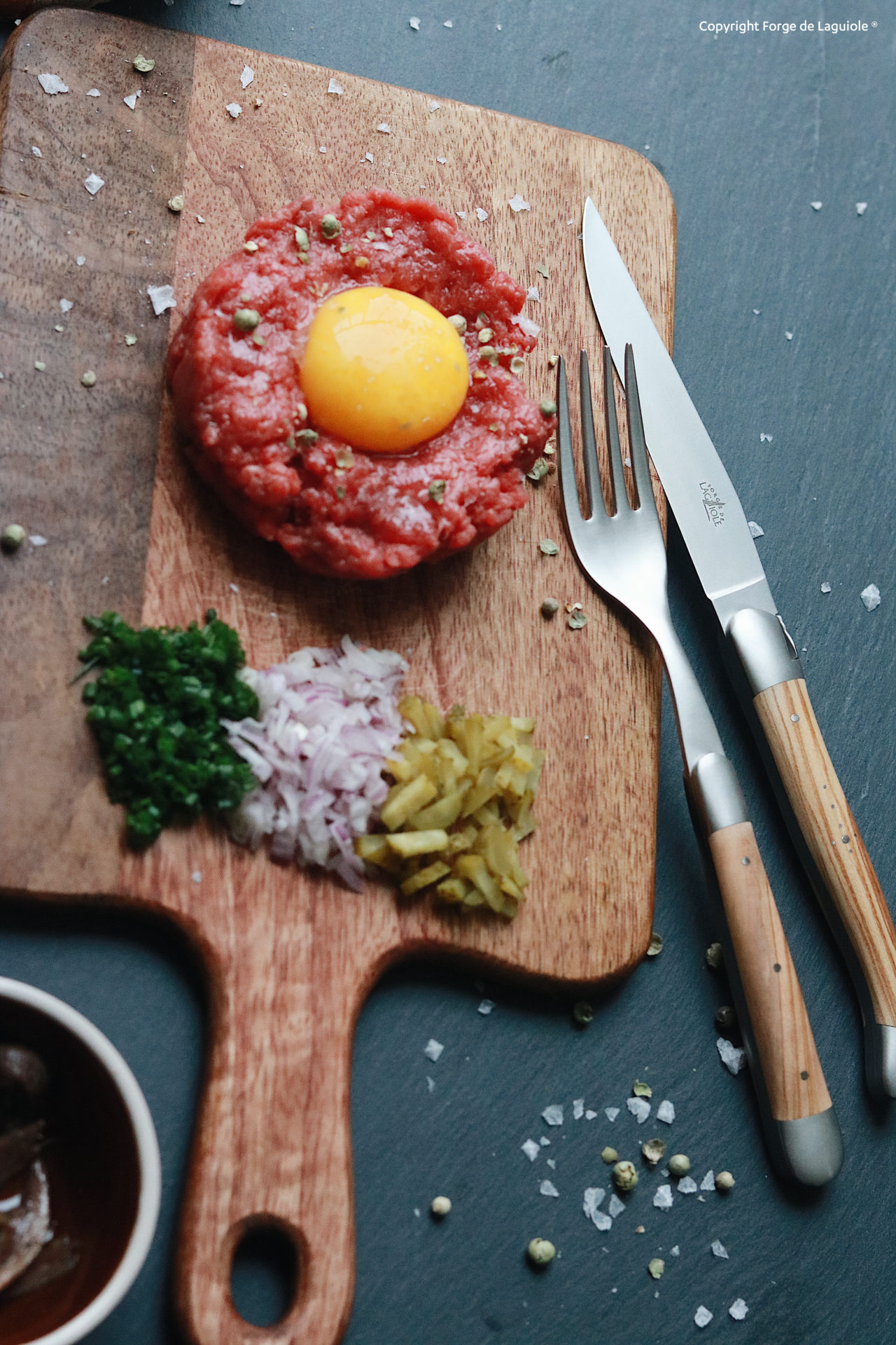Beef Tatar mit rohem Eigelb auf Holzbrett, daneben gehackte Zwiebel, Kräuter und Gewürzgurken, mit Besteck