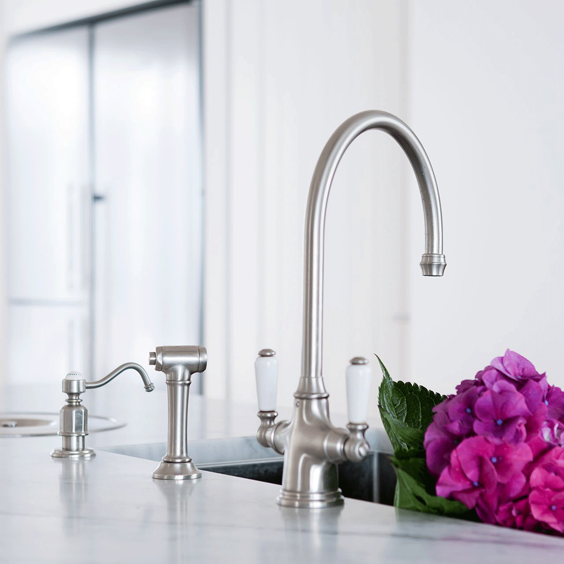 Moderne Edelstahl-Küchenarmatur mit klassischem Design an weißer Spüle, daneben lilafarbene Hortensienblüte