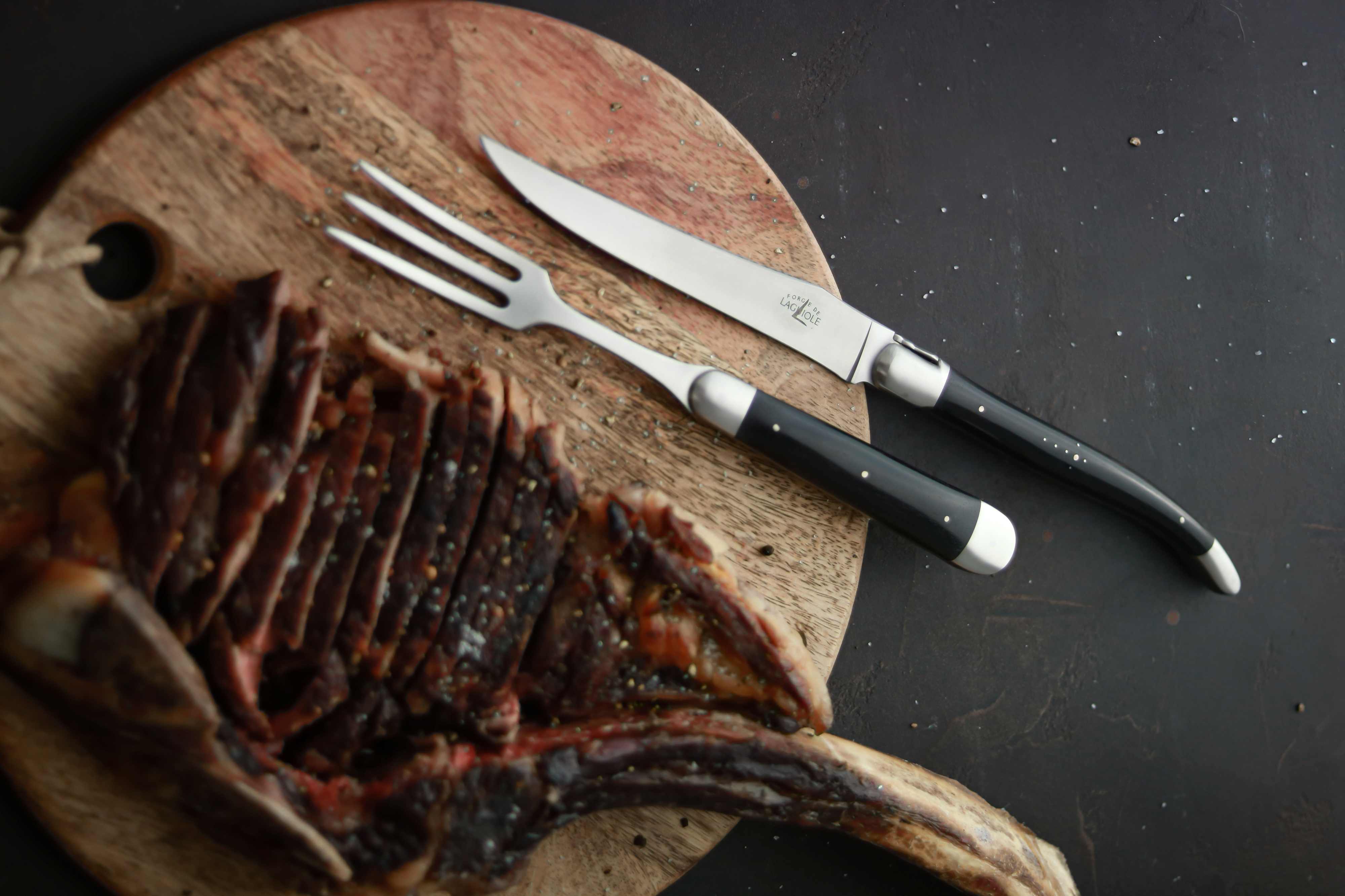 Ein Steakmesser und eine Fleischgabel mit schwarzem Griff liegen neben aufgeschnittenem, gewürztem Steak auf einem runden Holzbrett