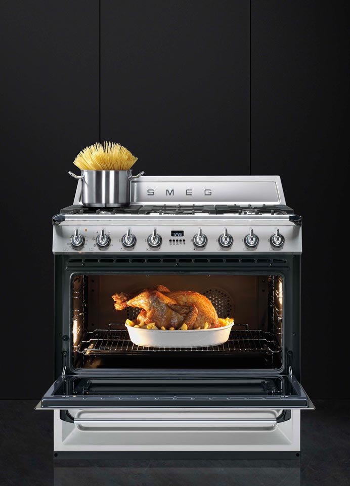 Moderner Smeg Standherd, auf dem Kochfeld steht ein Topf mit rohen Spaghetti, im geöffneten Ofen befindet sich ein goldbraun gebratenes Hähnchen in einer Auflaufform