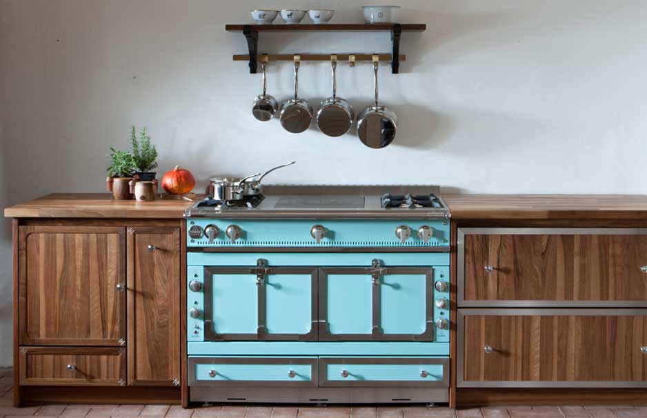 Küchenzeile mit türkisem Retro-Herd, Arbeitsplatte aus Holz, Hängeregal mit Töpfen und Teetassen, sowie Pflanzen und Gemüse auf der Seite