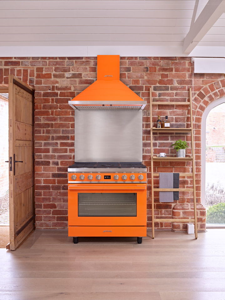 Orangefarbener Standherd mit passender Abzugshaube in einer Küche mit gemauerter Backsteinwand, Holzboden und seitlichem Regal.