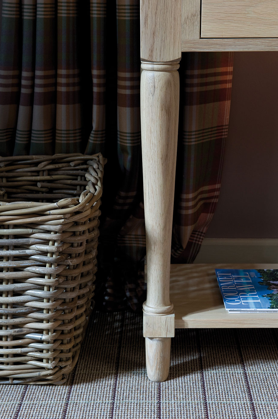 Nahaufnahme eines beigefarbenen Holztischbeins neben einem geflochtenen Korb, kariertem Vorhang und Magazin auf Teppich
