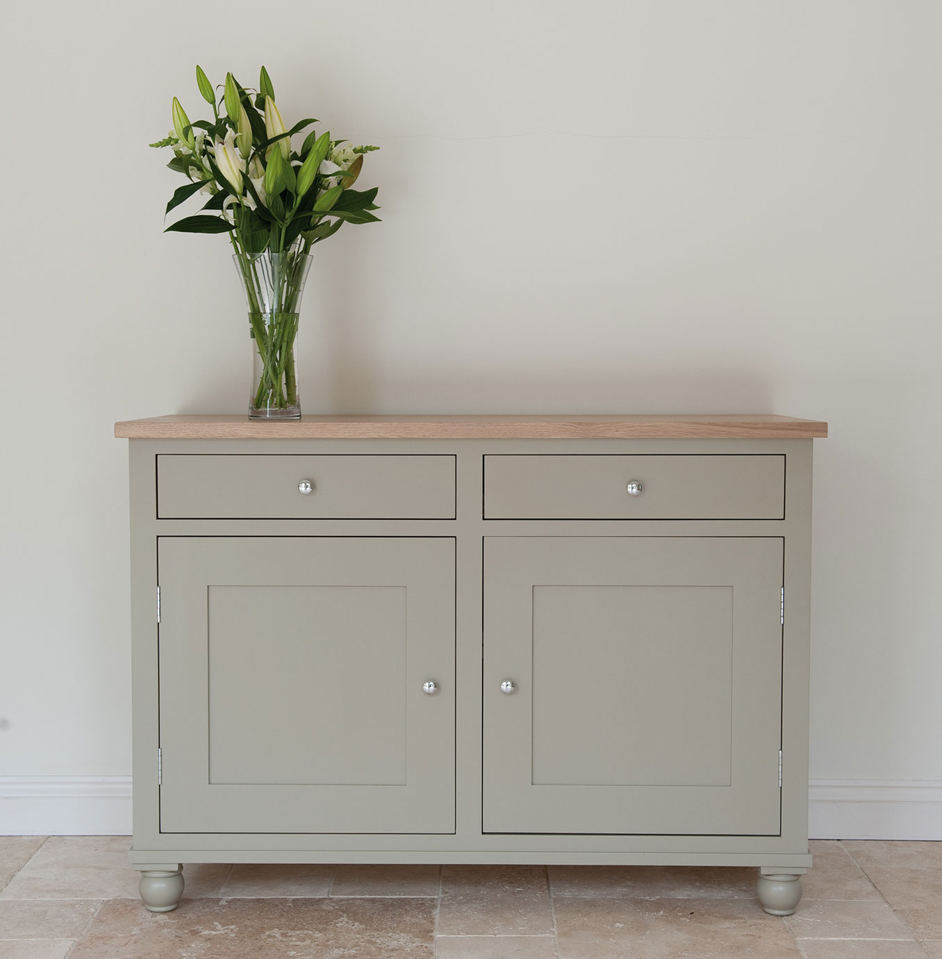 Helles Sideboard mit zwei Schubladen und zwei Türen, obere Platte aus Holz, darauf eine Glasvase mit weißen Lilien und grünen Blättern