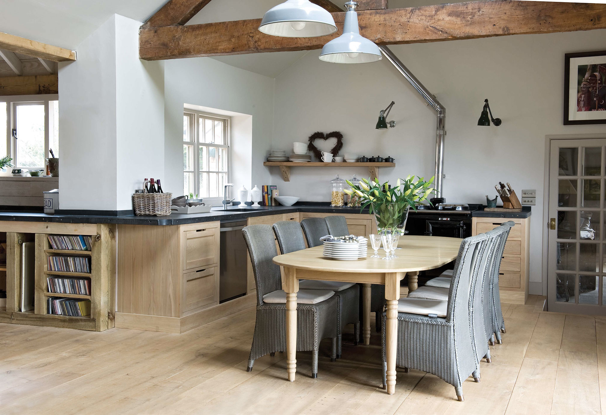 Helle Landhausküche mit Holzmöbeln, Esstisch, sechs geflochtenen Stühlen, dekorativen Pflanzen und offenen Regalen unter einem Holzbalken-Dach.