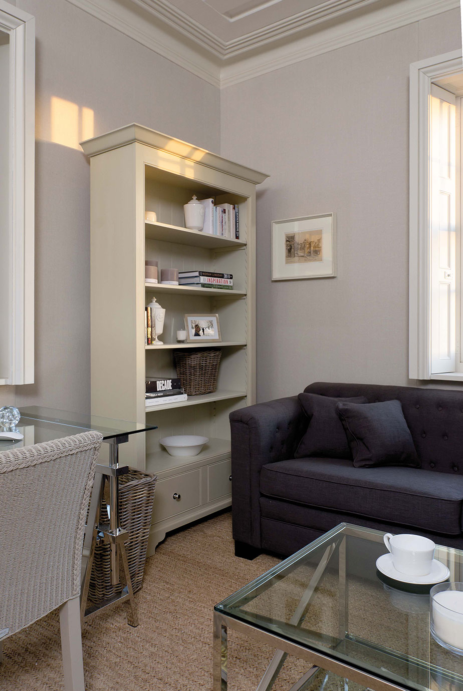 Modernes Wohnzimmer mit einem dunkelgrauen Sofa, Glas-Couchtisch, hellen Wänden, einem weißen Bücherregal und dekorativen Elementen für ein stilvolles Ambiente.