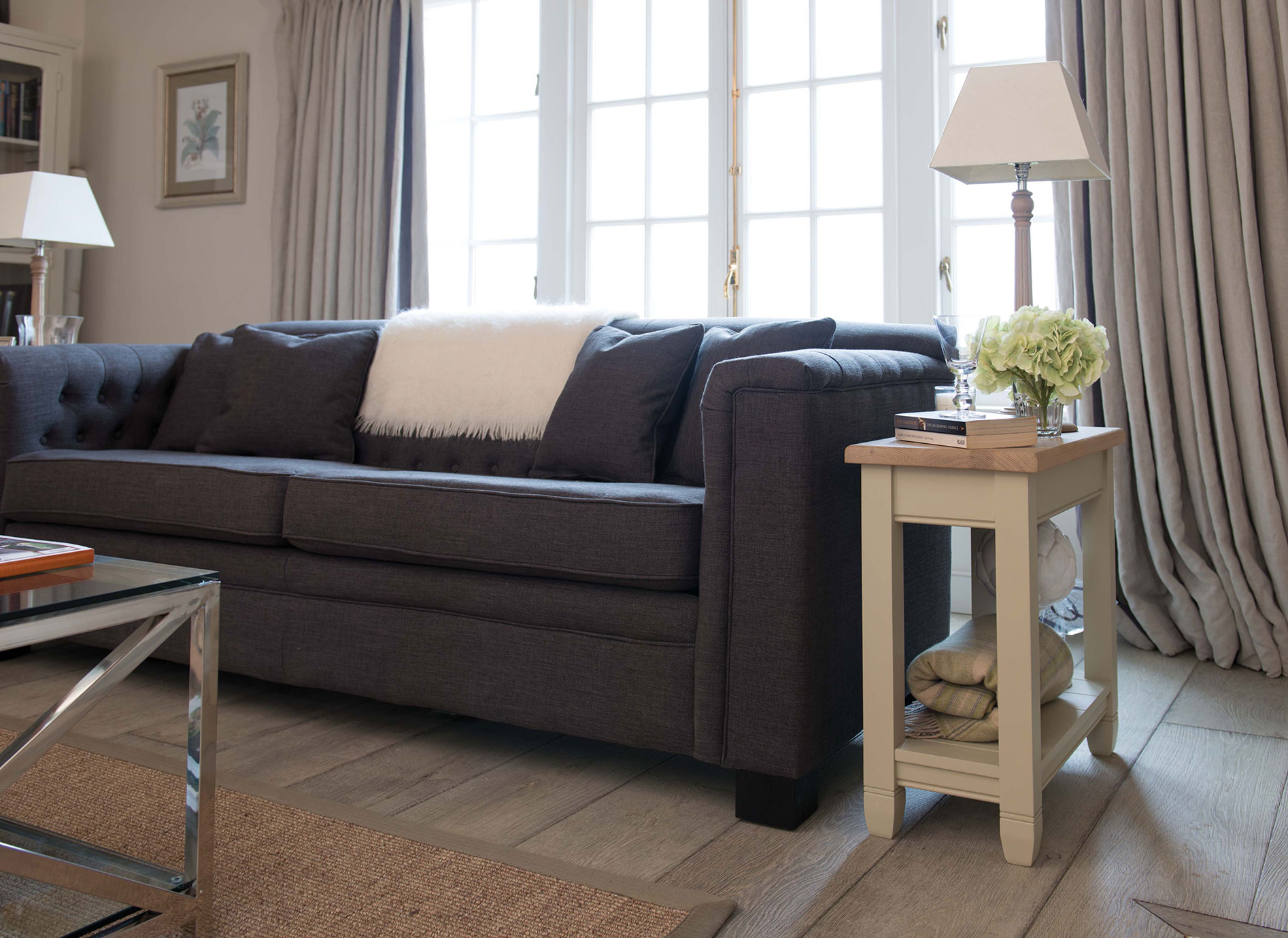 Gemütliches Wohnzimmer mit dunkelgrauem Sofa, Kissen, weißem Fell und Beistelltisch mit Lampe und Blumen