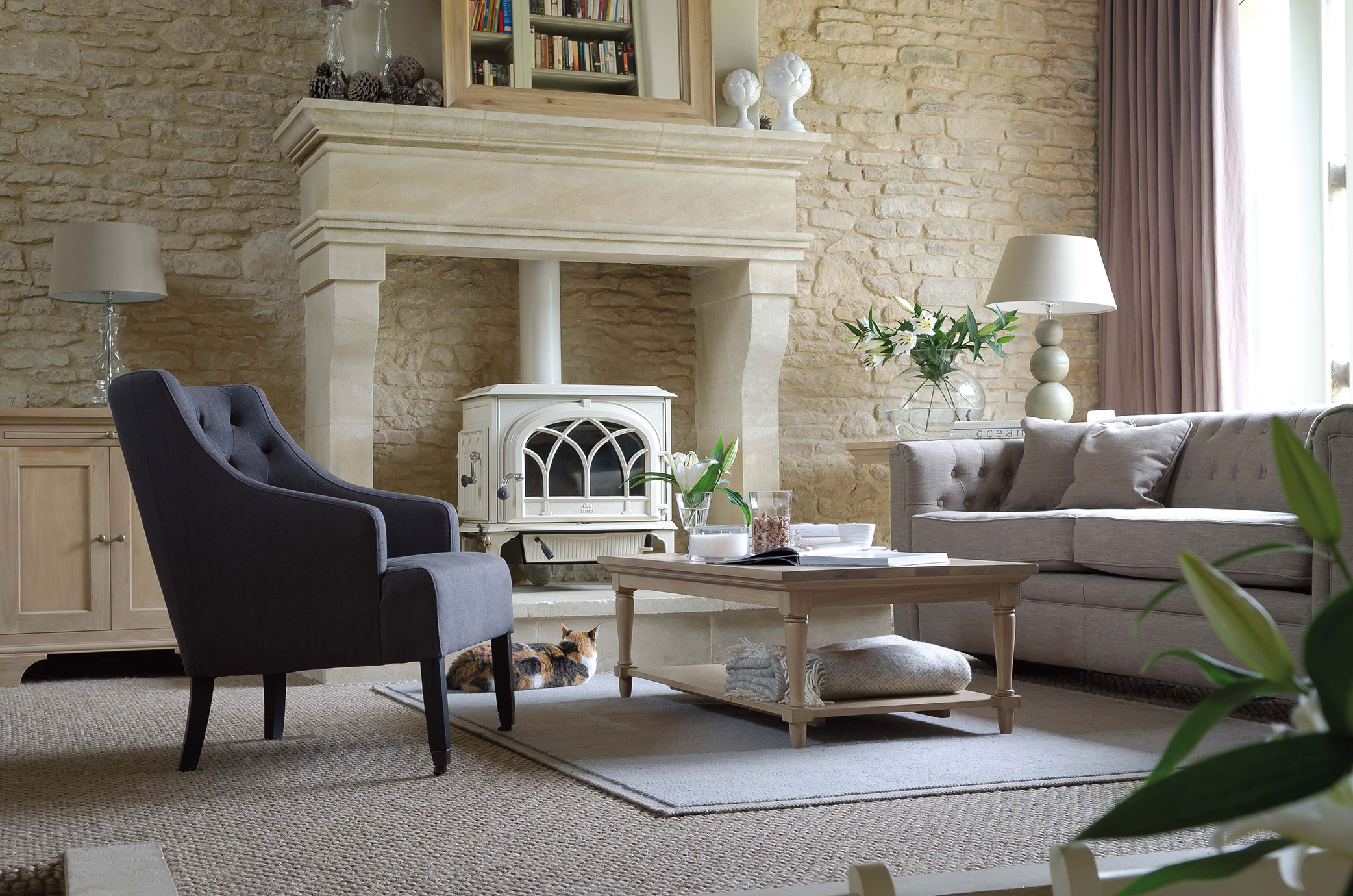 Helles Wohnzimmer mit steinerner Wand, Kaminofen, grauem Sessel, Sofa, Couchtisch, frischen Blumen und wohnlicher Atmosphäre