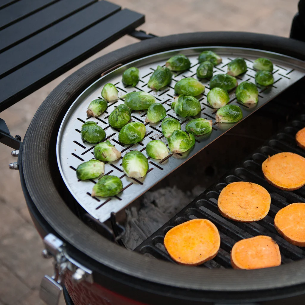 Rosenkohl auf einer Grillplatte und Süßkartoffelscheiben auf dem Grillrost eines runden Grills über Holzkohle