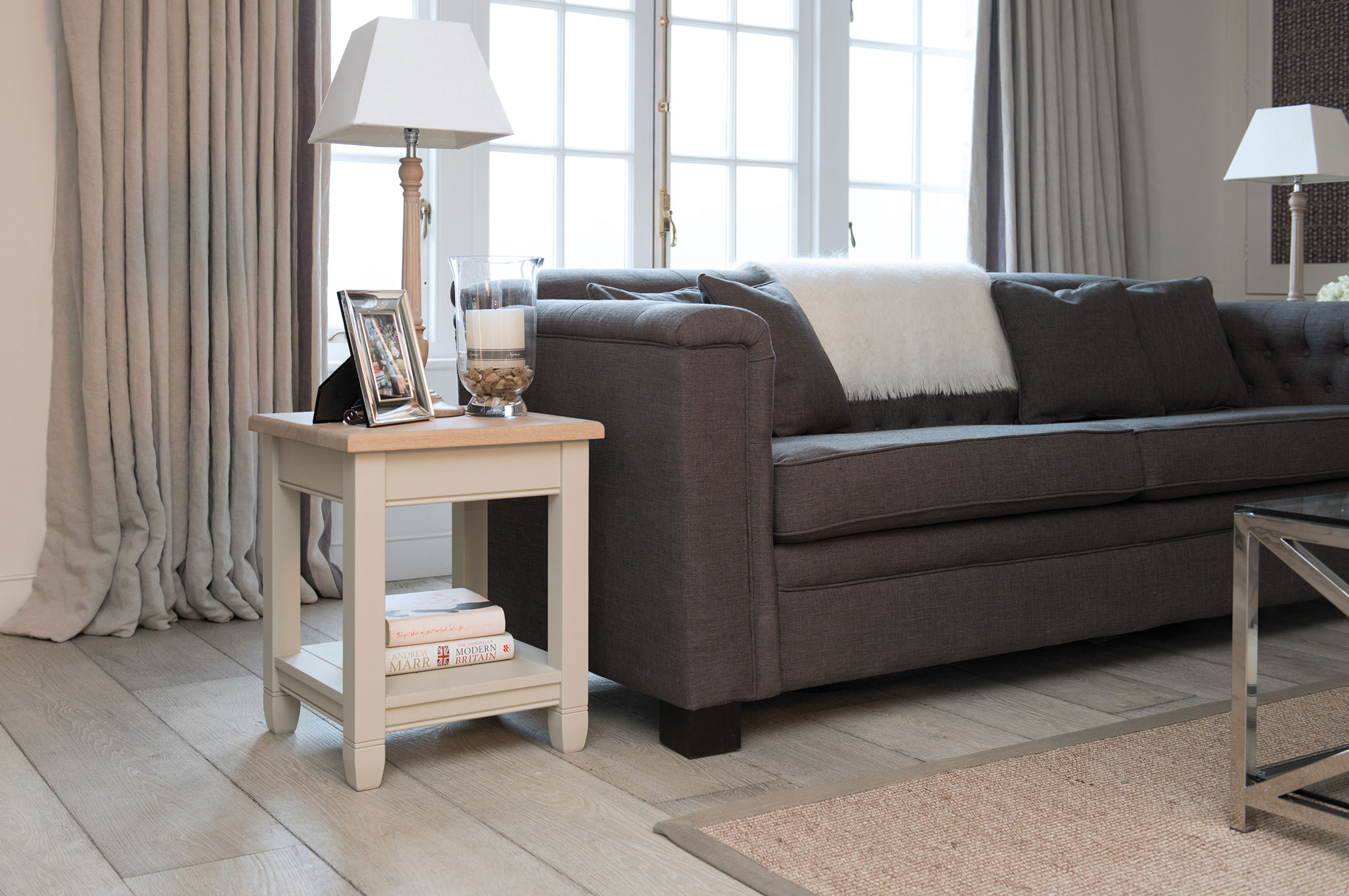 Gemütliches Wohnzimmer mit modernem grauen Sofa, beigem Beistelltisch, dekorativer Lampe, Büchern und Fensterfront