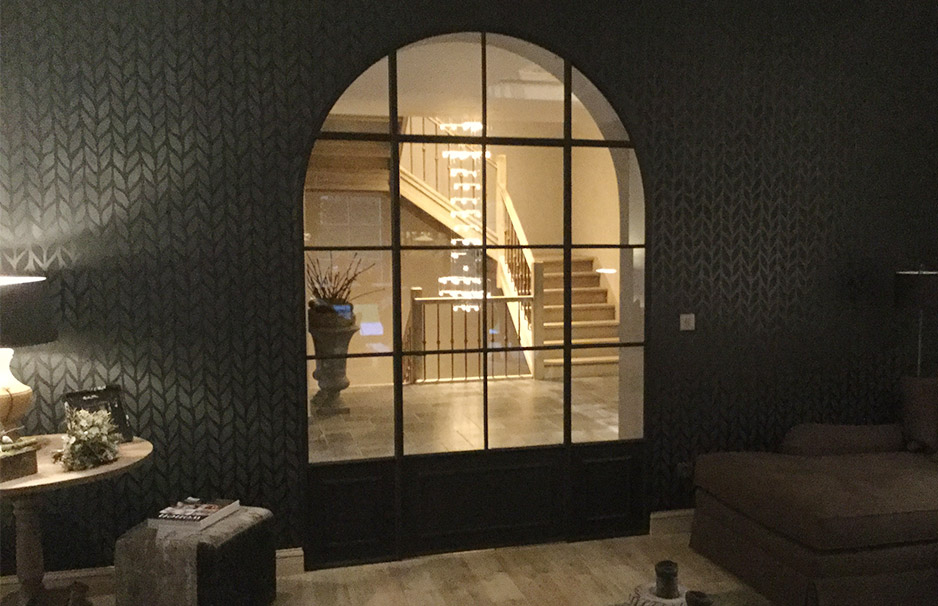 Schwarze Glaswand mit Rundbogen im Wohnzimmer, dahinter beleuchtetes Treppenhaus mit Holzgeländer und moderner Einrichtung