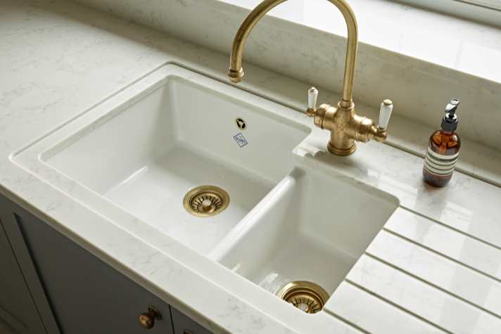 Elegantes weißes Keramik-Doppelspülbecken mit goldfarbenen Abflüssen und passender Küchenarmatur, daneben eine Seifenspenderflasche auf heller Arbeitsplatte