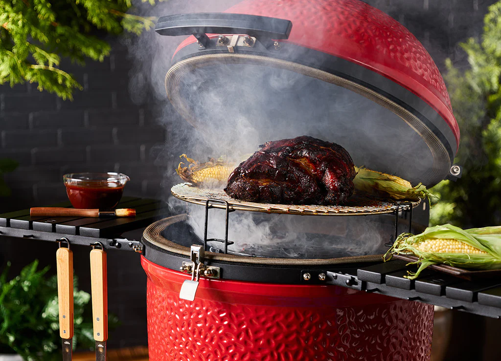 Ein roter Keramikgrill mit geöffnetem Deckel und rauchendem Grillrost, auf dem ein großes Stück Fleisch und Maiskolben liegen. Daneben steht eine Schüssel BBQ-Soße.