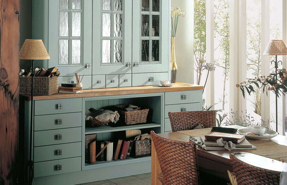 Esszimmer mit hellblauem Buffet, Glasvitrinen, Holztisch, geflochtenen Stühlen, natürlicher Beleuchtung und dekorativen Accessoires im Landhausstil.