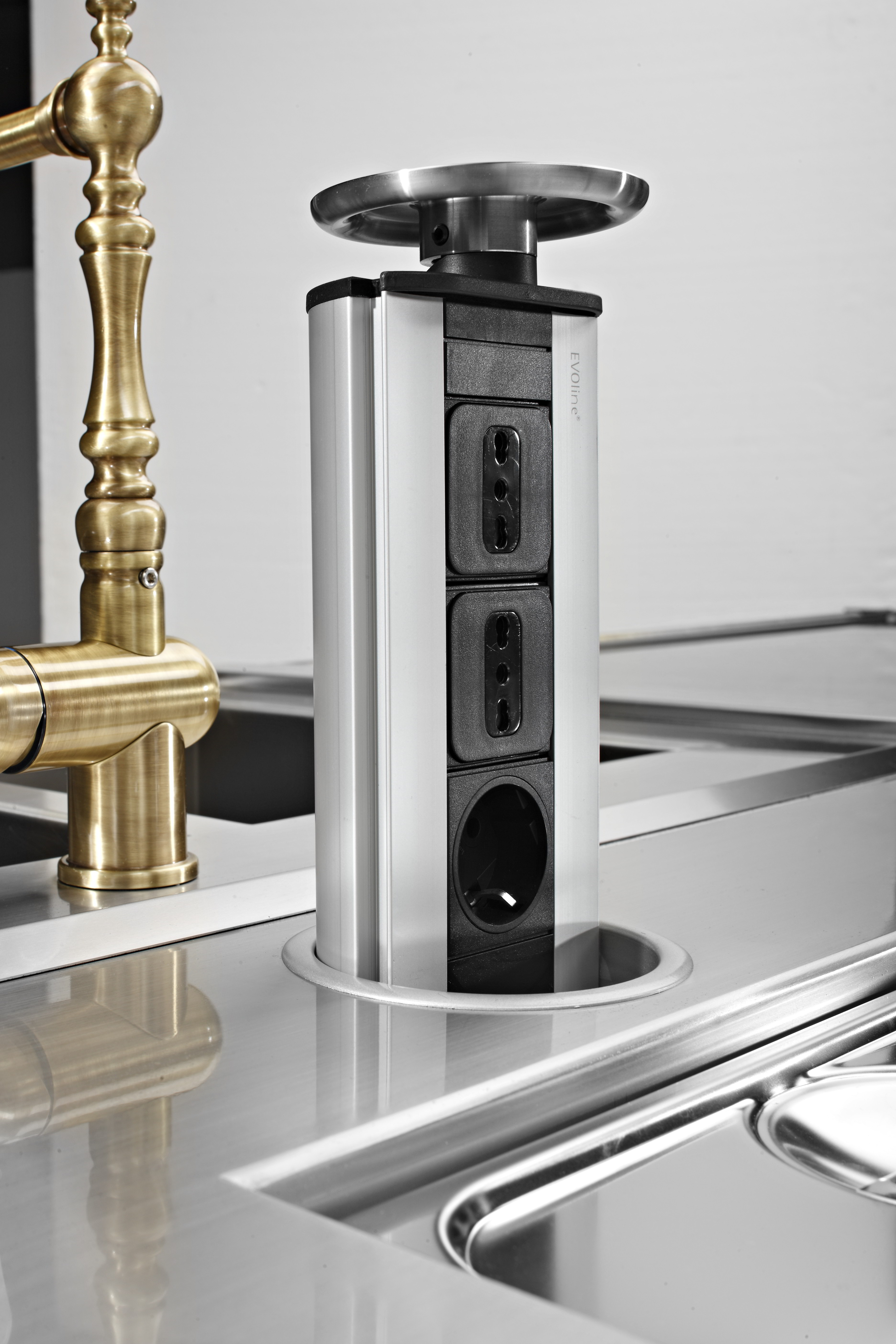 Hochwertige, versenkbare Steckdosenleiste aus Edelstahl und Kunststoff, eingebaut in eine Küchenarbeitsplatte neben einem goldfarbenen Wasserhahn