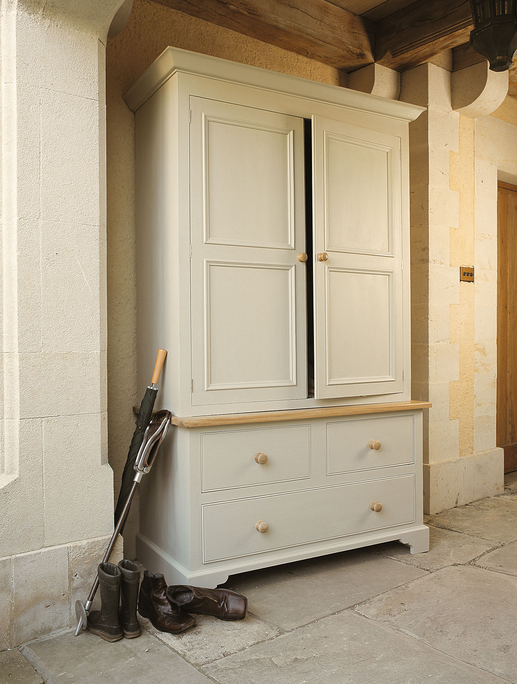 Eleganter Garderobenschrank aus Holz mit zwei Türen, zwei großen Schubladen und Holzgriffen. Davor stehen Regenschirm, Reitstiefel und Schuhe auf Steinboden.