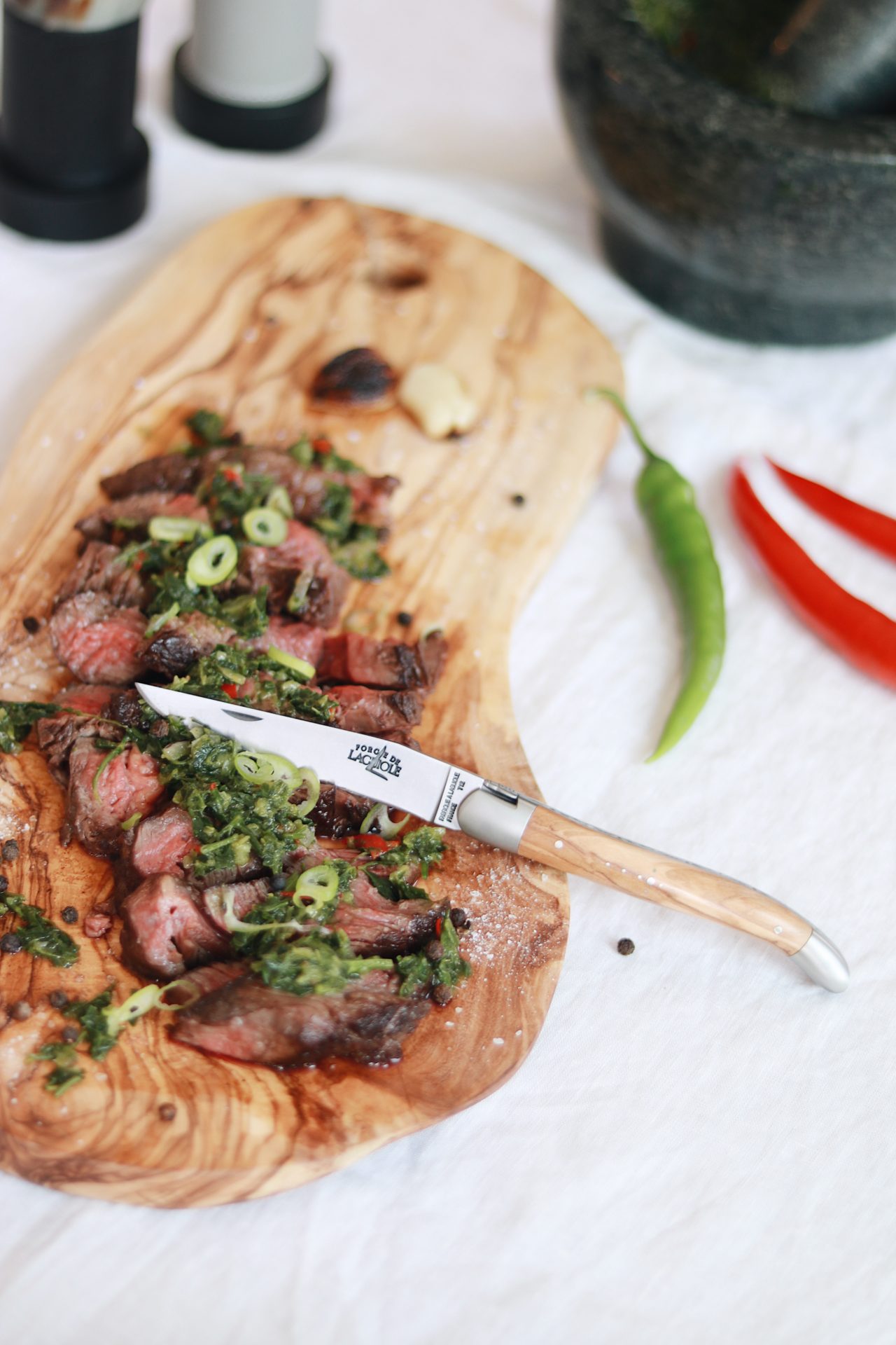 Laguiole-Messer mit Holzgriff auf Holzbrett, darauf saftiges Steak mit frischen Kräutern und Lauchzwiebeln angerichtet